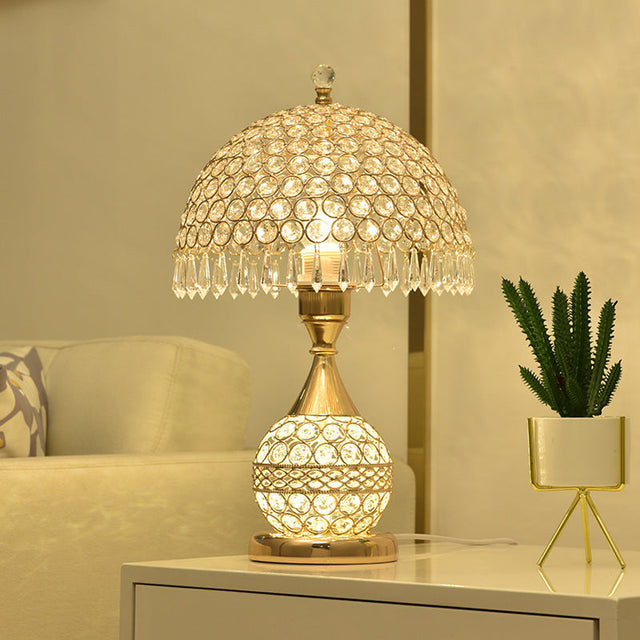 Gold crystal table lamp with a beaded shade lit on a bedside cabinet