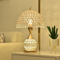 Gold crystal table lamp with a beaded shade lit on a bedside cabinet