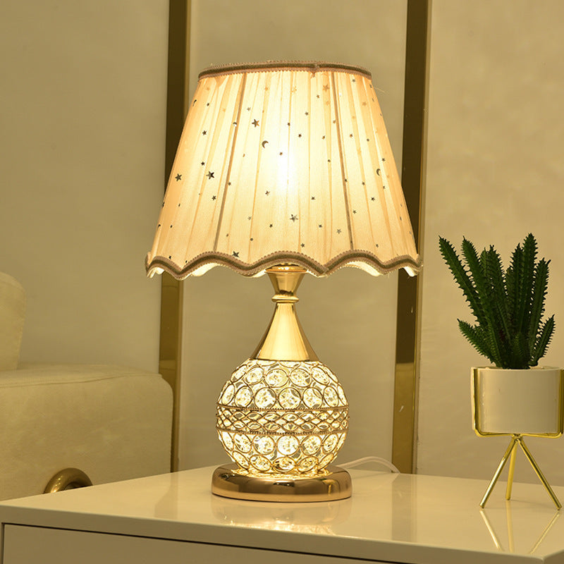 Gold crystal table lamp with a scalloped fabric shade lit on a cabinet