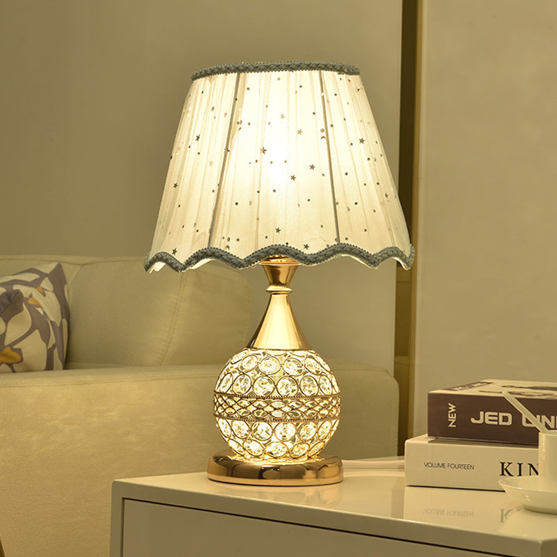 Gold crystal table lamp with a scalloped shade lit on a white cabinet