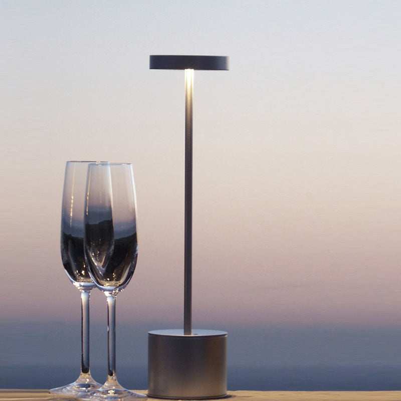 Black cordless table lamp lit beside two champagne glasses at dusk