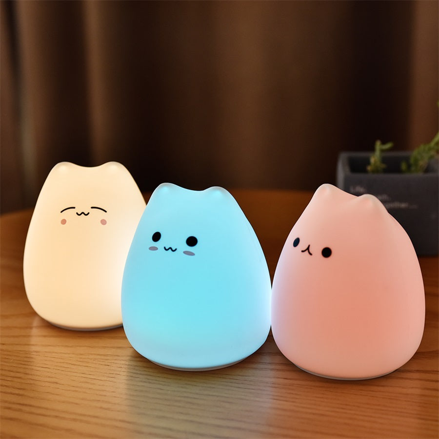 Three silicone cat night lights lit pink, blue and pale pink on a table