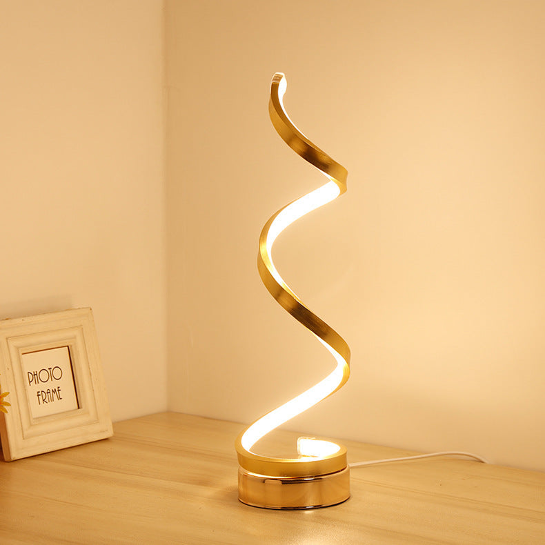 Gold spiral LED table lamp lit warm in a corner beside a picture frame