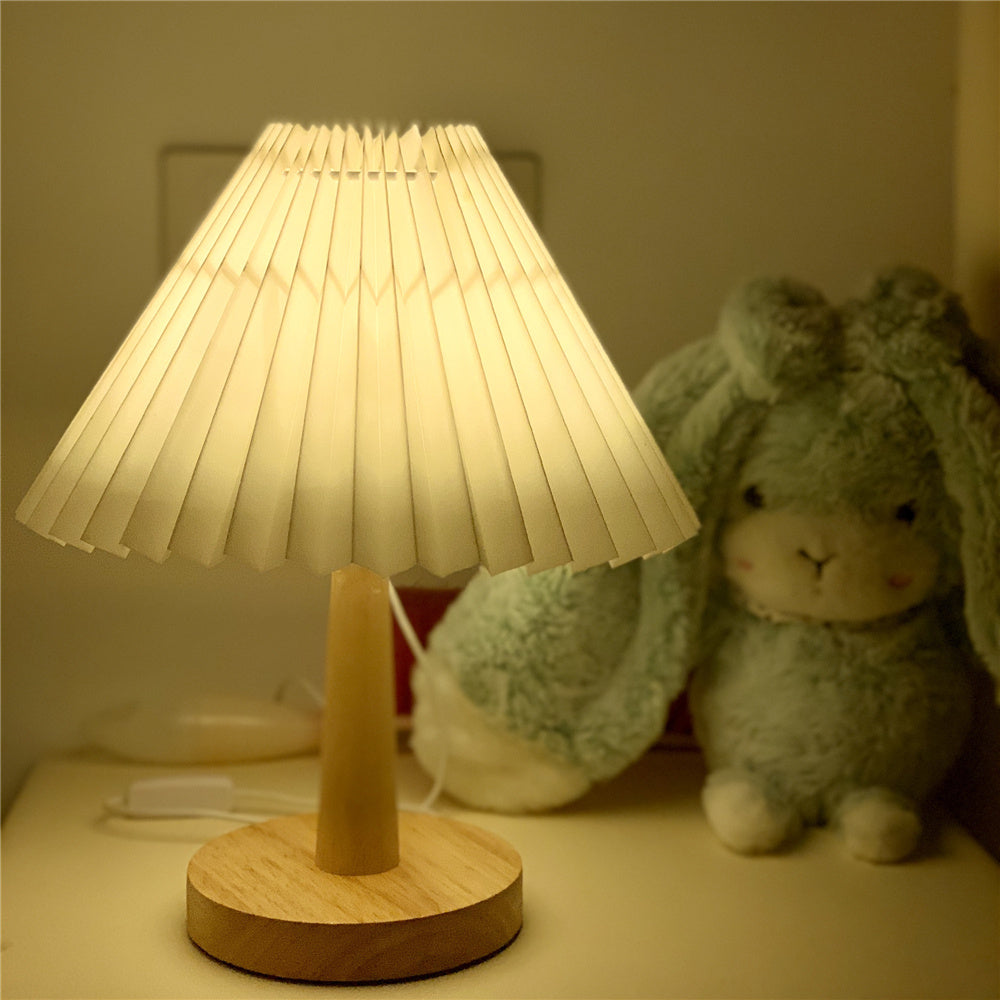 Pleated-shade table lamp lit warm on a nightstand beside a soft toy