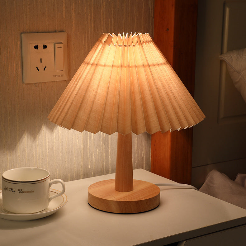 Beige pleated-shade table lamp lit on a bedside table beside a cup