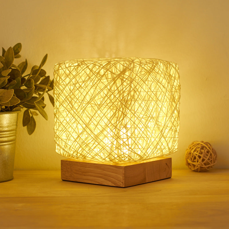 Woven rattan table lamp lit warm on a wooden base beside a plant