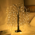 LED willow tree light with warm drooping branches glowing on a bed